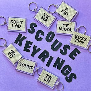 May include: A collection of clear acrylic keychains with embroidered designs and silver keyrings. Each keychain features a different phrase in black thread on a beige background. The words "SOFT LAD", "YE MA", "R KID", "YE WOOL", "BOSS THA", "MG DO", "SOUND", and "TA LA" are visible.