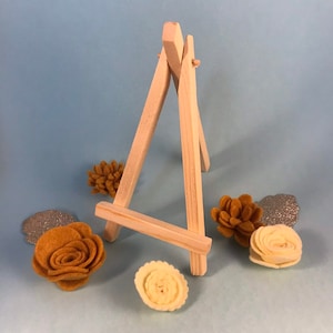 Könnte beinhalten: Ein kleiner Holzstaffelei, perfekt zum Ausstellen von kleinen Kunstwerken oder Fotos. Die Staffelei ist aus Naturholz gefertigt und hat ein schlichtes, rustikales Design. Sie ist von mehreren Filzblumen in Gelb- und Brauntönen umgeben.