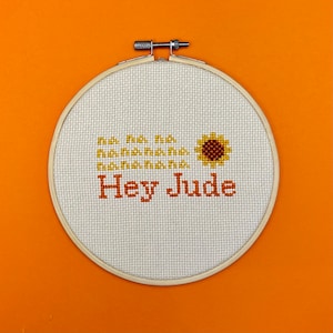 May include: A white embroidery hoop with a cross-stitch design of a sunflower and the text "Hey Jude".