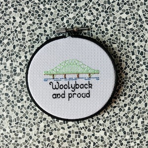 Può includere: Un cerchio da ricamo bianco con un disegno di ponte verde e il testo "Woolyback and proud".
