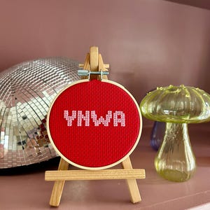 May include: A small, round cross-stitch embroidery hoop with the letters "YNWA" in pink on a red background. The hoop is displayed on a miniature wooden easel. A disco ball and a glass mushroom are in the background.