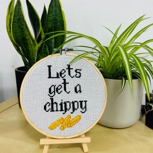 May include: A circular wooden embroidery hoop containing white Aida fabric with black cross-stitched text that reads "Lets get a chippy" above a small yellow cross-stitched graphic of three chips, displayed on a small wooden easel stand.