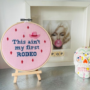 May include: A pink cross-stitch hoop with the text "This ain't my first RODEO" and a cowboy hat design. The hoop is displayed on a small wooden easel. A framed Marilyn Monroe print and a decorative skull are in the background.