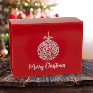 May include: A festive red gift box featuring a white Christmas bauble design and the words "Merry Christmas". The box is placed on a wooden surface, with a blurred Christmas tree in the background.