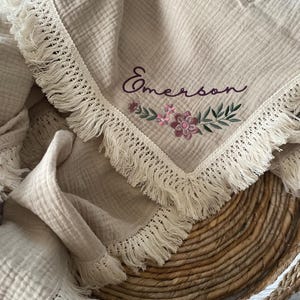 May include: A beige muslin baby blanket with a white crocheted trim and fringe. The blanket is embroidered with the name "Emerson" and a floral design in pink and purple.