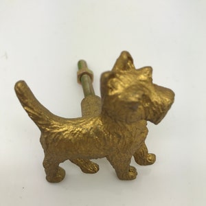 May include: Gold metal cabinet knob shaped like a terrier dog. The dog is standing on all four legs with its tail curled up. The knob has a screw on the back.
