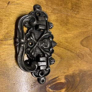 May include: A black metal drawer pull with a skull and crossbones design. The pull has a decorative scrollwork design and is attached to a wooden surface.