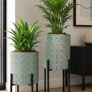 May include: Two cylindrical planters with a teal and gold fan-shaped embossed pattern, each supported by a black metal stand with four thin legs. Each planter holds a green leafy plant with long, pointed foliage, displayed at different heights.