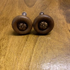 May include: Two brown wooden cabinet knobs with a gold flower design in the centre.