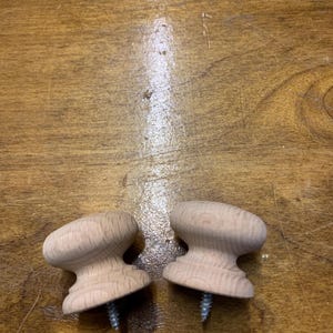 May include: Two unfinished wooden cabinet knobs with a mushroom-shaped design. Each knob has a screw for easy installation. The knobs are made of light-coloured wood and are ready to be painted or stained.