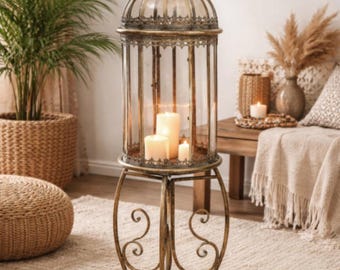 May include: A decorative gold-colored metal and glass candle lantern with three lit candles inside. The lantern has an ornate, cage-like design and stands on a three-legged base with scrolled details. The lantern is approximately 30 inches tall.