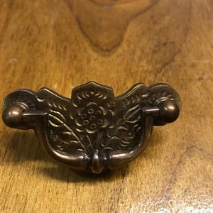 May include: A brass cabinet pull with a floral design. The pull has a curved shape and two knobs on the sides.