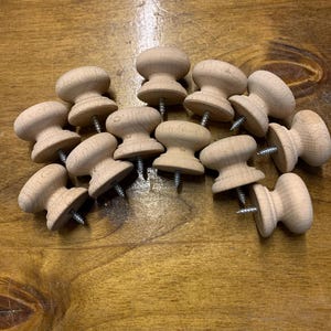 May include: A collection of unfinished wooden knobs with attached screws. The knobs have a mushroom-like shape, with a rounded top and a wider base. The wood has a light, natural colour. The knobs are scattered on a wooden surface.