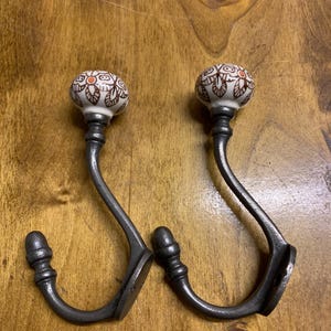 May include: Two decorative hooks with a white ceramic knob featuring brown floral designs. The hooks are made of metal and have a dark grey finish.