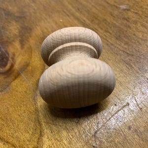 May include: A turned wooden furniture knob, featuring a rounded top and base joined by a narrower, cylindrical midsection. The unfinished wood grain is visible, displaying the natural colour and texture. This knob is likely for a cabinet or drawer.