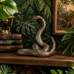May include: A bronze-coloured rattlesnake figurine with a textured, scaled surface. The snake is coiled with its head raised and its rattle pointed upwards. The sculpture features intricate detailing on the scales and a realistic, alert posture.