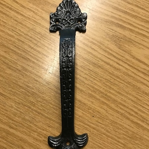 May include: Black cast iron door handle with ornate details. The handle has a pointed top and bottom with a decorative design.