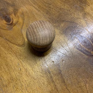 May include: A small, round wooden knob in a light brown hue. The knob features a circular top and a smaller base. The wood grain is clearly visible on the top surface. This knob is likely for a drawer or cabinet.