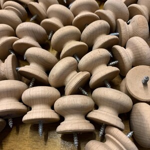May include: A pile of unfinished wooden knobs with screws attached. The knobs are mushroom-shaped with a flat base. The wood grain is visible, and the colour is a light, natural tone. Each knob has a screw for easy installation.