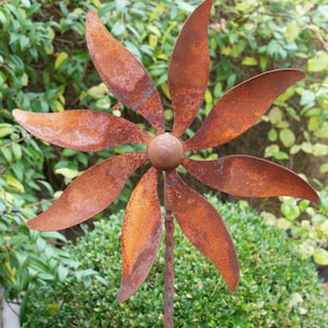 May include: A weathered metal garden sculpture in the shape of a flower. The flower features seven rusty brown petal-shaped blades around a central sphere. The stem is a twisted metal rod. The background consists of green foliage.