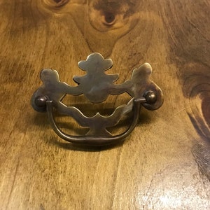 May include: A brass drawer pull with a decorative, ornate design. The pull has a curved, D-shaped handle and a floral-inspired top.
