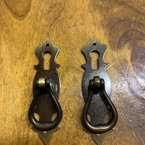 May include: Two antique brass drawer pulls. Each pull features a decorative backplate with a keyhole shape and a curved handle. The pulls are a dark brass colour and are set against a wooden surface.