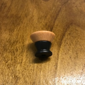May include: A wooden knob with a black base. The knob is light brown and has a smooth, rounded top.