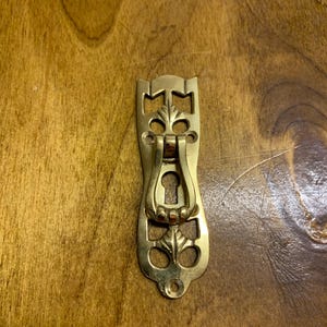 May include: A brass door pull with a keyhole and intricate floral designs. The pull is in the shape of a teardrop and has a rounded top.