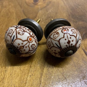 May include: A pair of decorative cabinet knobs. Each knob has a white ceramic sphere with a brown floral pattern and orange accents. The knobs have a dark metal base and a screw for attachment, suitable for furniture or decor.
