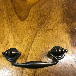 May include: A dark brown wooden surface with a decorative, antique-style, black metal drawer pull. The pull has a curved, elongated shape with a rounded knob on each end.