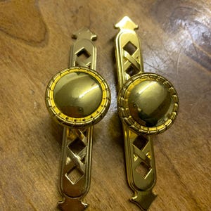 May include: Two gold-coloured cabinet pulls. Each pull has a round, ridged knob and an elongated backplate with a diamond-shaped lattice design. The pulls are made of a shiny, metallic material.