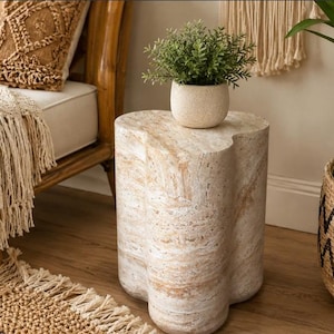 May include: A clover-shaped side table crafted from light beige and cream-coloured stone with a natural marbled texture. The table features a smooth, flat top surface and a solid, continuous base, supporting a small, round, speckled beige planter.