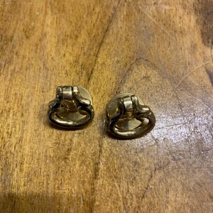 May include: A pair of antique brass drawer pulls, each featuring a ring handle. The circular pulls have a decorative base and a small ring attached. The brass has an aged appearance, indicating vintage or antique hardware.