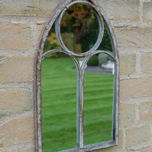 May include: An arched wall mirror with a weathered, distressed metal frame. The design features a pointed gothic-style top with a circular upper section and two rectangular lower panes, reflecting a green lawn and trees in the glass surface.