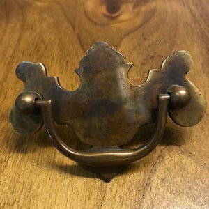 May include: Vintage brass drawer pull with a decorative backplate and a curved handle. The pull has a tarnished finish, showing signs of age. The backplate features an ornate, symmetrical design with rounded edges and a central decorative element.