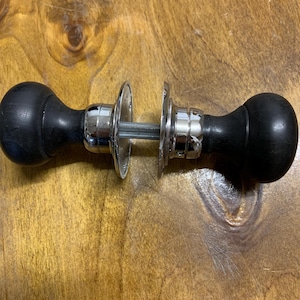 May include: A black door knob set with a polished chrome finish. The door knobs are round and smooth, with a classic design. The set includes two knobs and a connecting rod, suitable for interior doors.