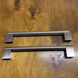 May include: Two brushed nickel cabinet pulls with a rectangular shape. The pulls are on a wooden surface.