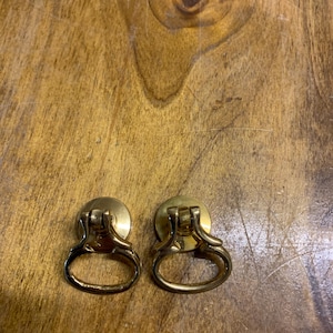 May include: A pair of antique brass drawer pulls, each featuring a ring handle. The pulls have a circular base and a curved handle, ideal for vintage furniture. The brass has a warm, golden colour.