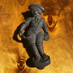May include: A cast iron figurine of a person wearing a hat and holding a small object. The figurine is painted black and is standing on a round base.