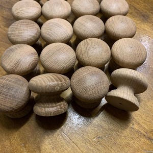 May include: Twenty-one unfinished wooden knobs are displayed on a wooden surface. The knobs are round with a textured surface, and some have a mushroom-shaped design. The wood has a natural, light brown colour.
