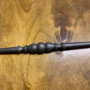 May include: A dark-finished wrought iron decorative rod. The rod has textured, spherical embellishments in the centre, and tapers at each end. The rod is designed for decorative use, possibly for a curtain or similar.