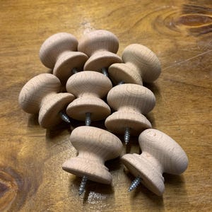 May include: A pile of unfinished wooden cabinet knobs with screws attached. The knobs have a rounded top and a flared base, displaying the natural wood grain. Suitable for furniture restoration or new projects.