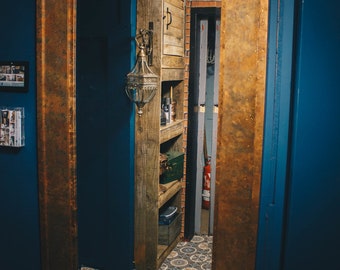 Op de afbeelding: Een koperkleurige spiegel met lijst reflecteert een kamer met een houten rek en een betegelde vloer met een patroon. Het rek heeft een lamp en verschillende items op de planken.