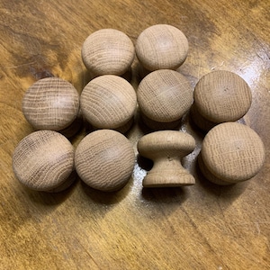 May include: A collection of wooden cabinet knobs and a single wooden cabinet pull. The knobs are round with a natural wood grain finish. The cabinet pull has a mushroom-like shape. The items are arranged on a wooden surface.