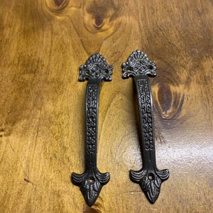 May include: Two black metal door handles with ornate designs. The handles feature a decorative floral pattern and a pointed tip.