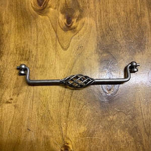 May include: A dark metal drawer pull with a central woven design. The handle has a curved shape with ball-shaped ends. The metal has a rustic, aged appearance, suggesting a vintage or antique style. Suitable for furniture restoration or adding a unique touch.