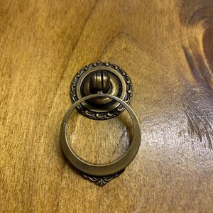 May include: A brass ring pull cabinet door handle with a decorative, ornate, circular design.