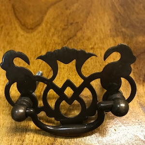 May include: A black metal drawer pull with a decorative, ornate design. The pull has two round knobs on either side and a curved handle in the centre.