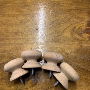 May include: Four unfinished wooden cabinet knobs with attached screws are arranged on a wooden surface. The knobs have a mushroom-like shape, with a rounded top and a wider base. The wood grain is visible, and the knobs appear ready for staining or painting.