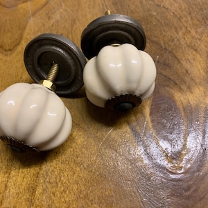 May include: Four cream-coloured, pumpkin-shaped cabinet knobs with metal hardware are shown. The knobs have a ribbed design and are set against a wooden surface. The metal hardware is a dark grey colour.